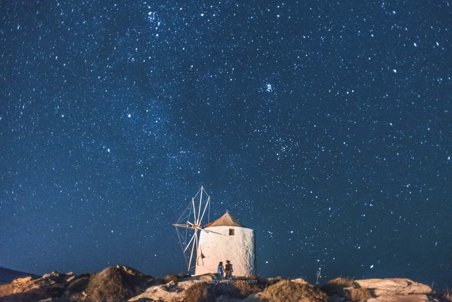 Couple standing beside a traditional Greek windmill under a starry sky photographed by KrystaPhotos, winner of 2nd place at the Greeka Greek Islands photo contest