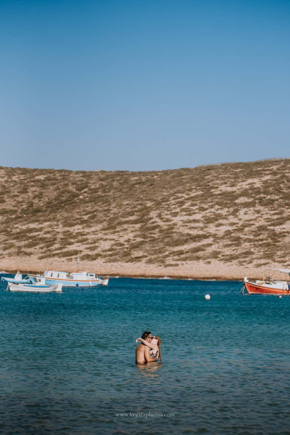 Couple embracing in the sea with fishing boats and hills in the background, Amorgos, Greece