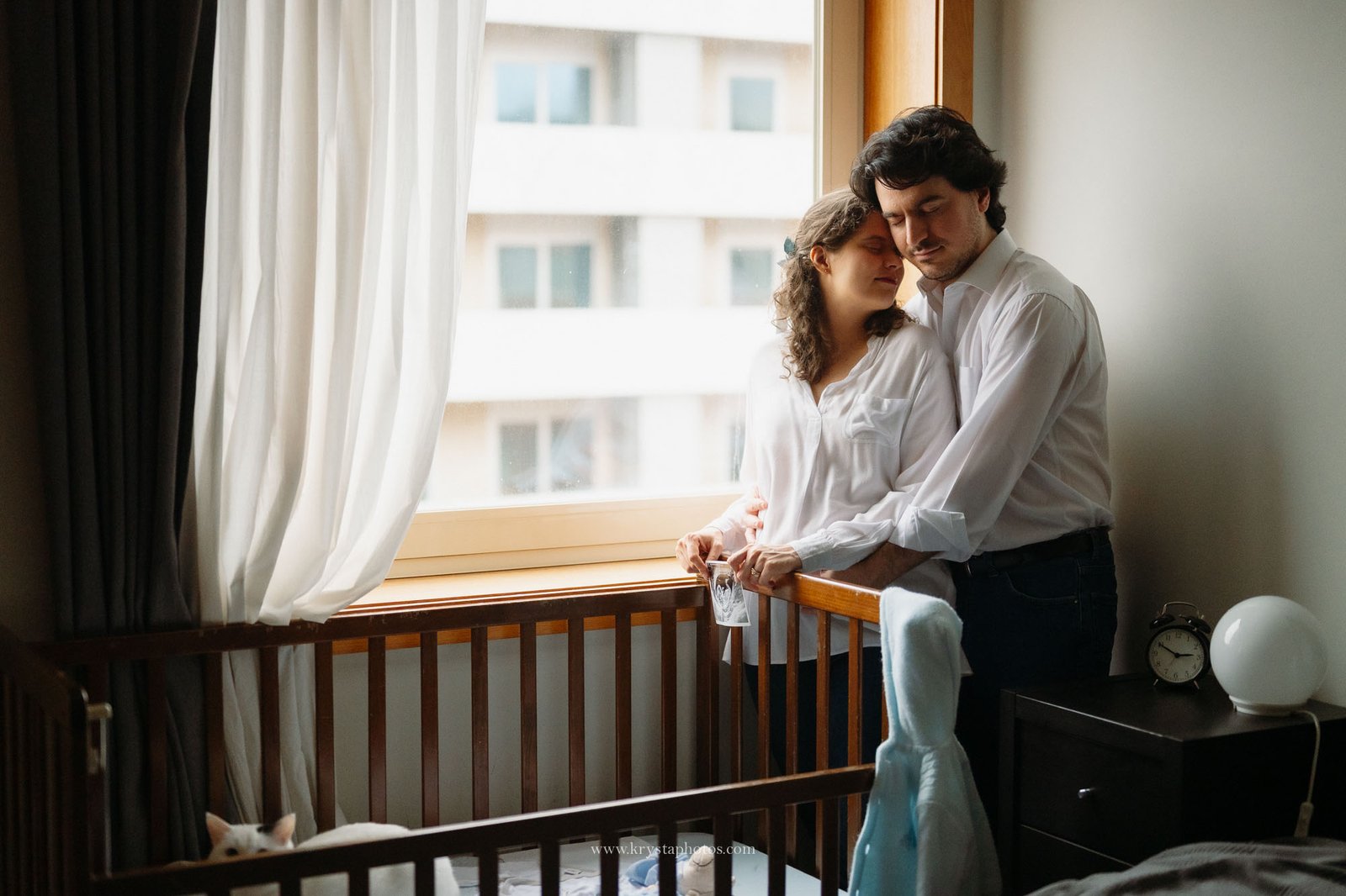 Expecting couple standing together beside a baby crib in their home, embracing softly near a window with natural light during an intimate at-home maternity photography session.