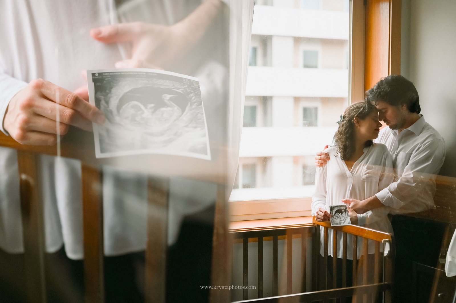Expecting couple standing together beside a baby crib in their home, embracing softly near a window with natural light during an intimate at-home maternity photography session.