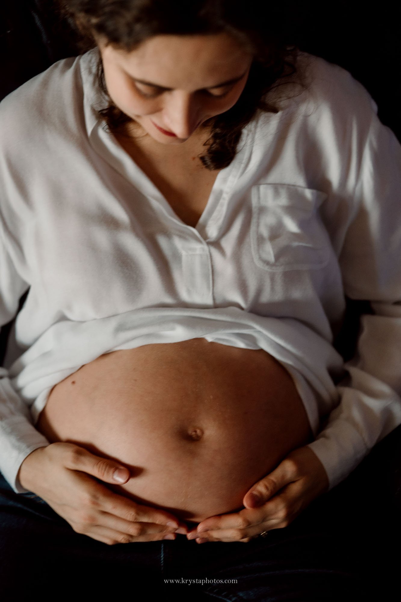 Expecting couple standing together beside a baby crib in their home, embracing softly near a window with natural light during an intimate at-home maternity photography session.