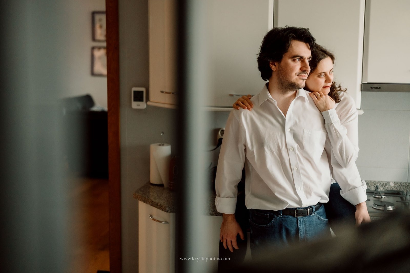 Expecting couple standing together beside a baby crib in their home, embracing softly near a window with natural light during an intimate at-home maternity photography session.