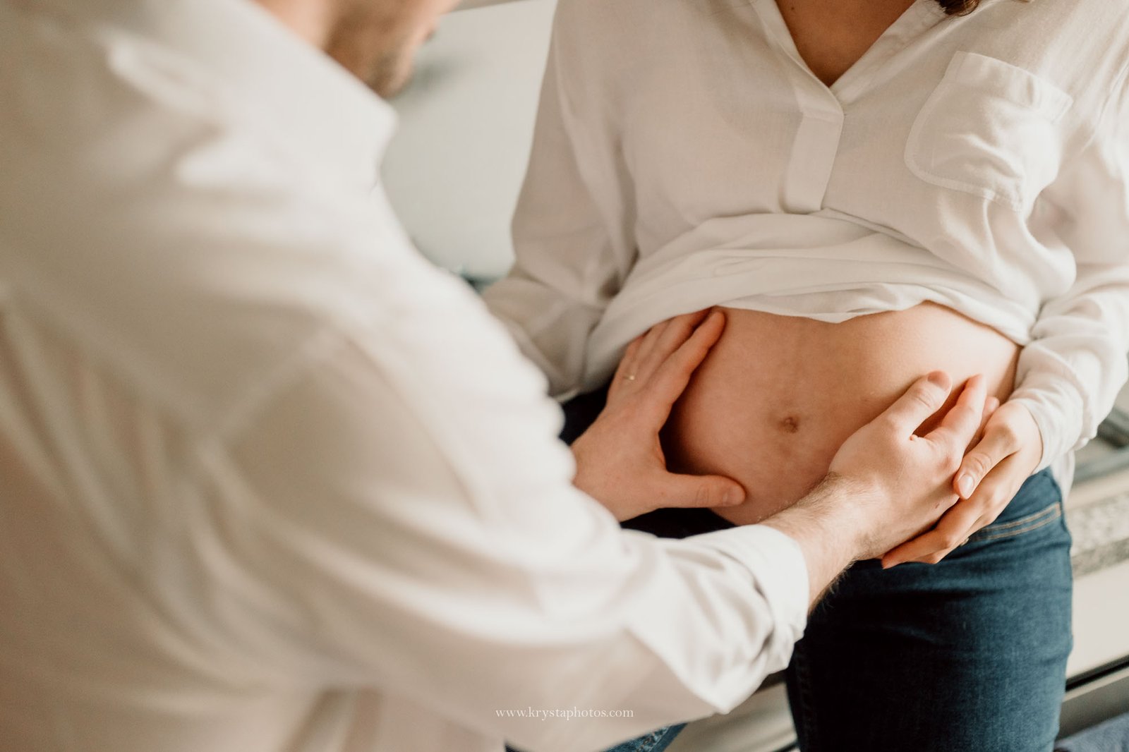 Close up maternity photo of an expecting couple embracing in their kitchen, with the mother’s hand resting gently on the partner’s shoulder, captured indoors using natural light in a documentary style.