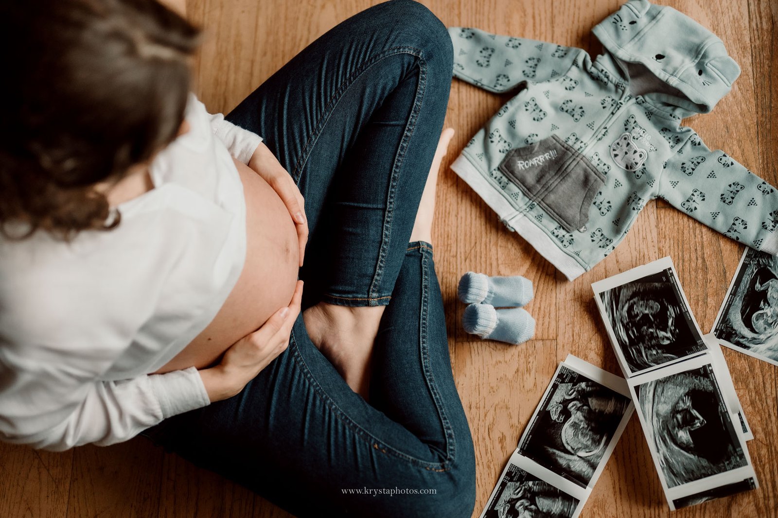 Top view maternity photo of an expecting mother sitting on the floor, holding her belly, surrounded by baby clothes, tiny socks, and ultrasound photos, captured indoors using natural light.
