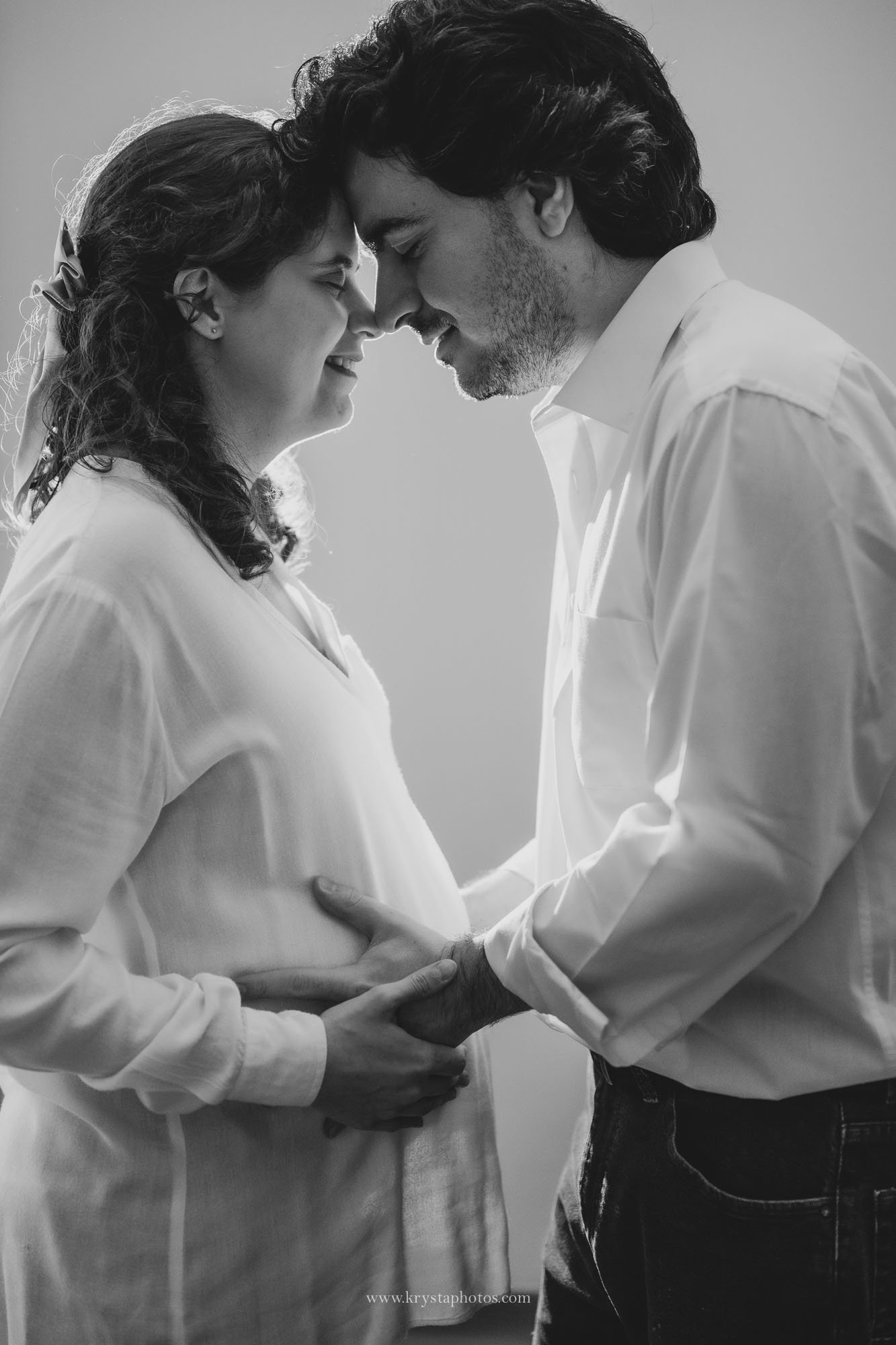 Expecting couple standing together beside a baby crib in their home, embracing softly near a window with natural light during an intimate at-home maternity photography session.
