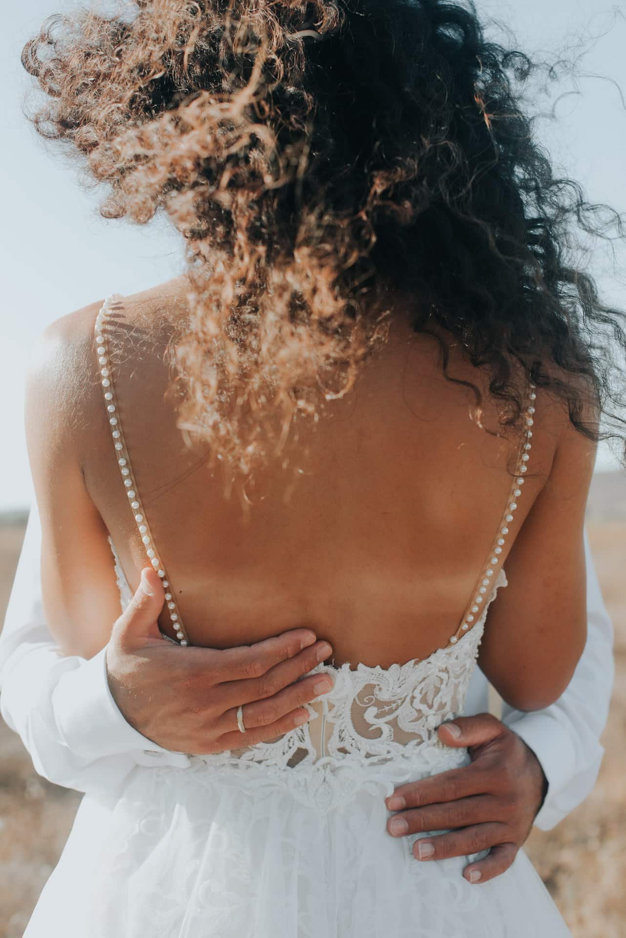 Groom embracing bride from behind showing low back pearl strap wedding dress detail on Greek beach