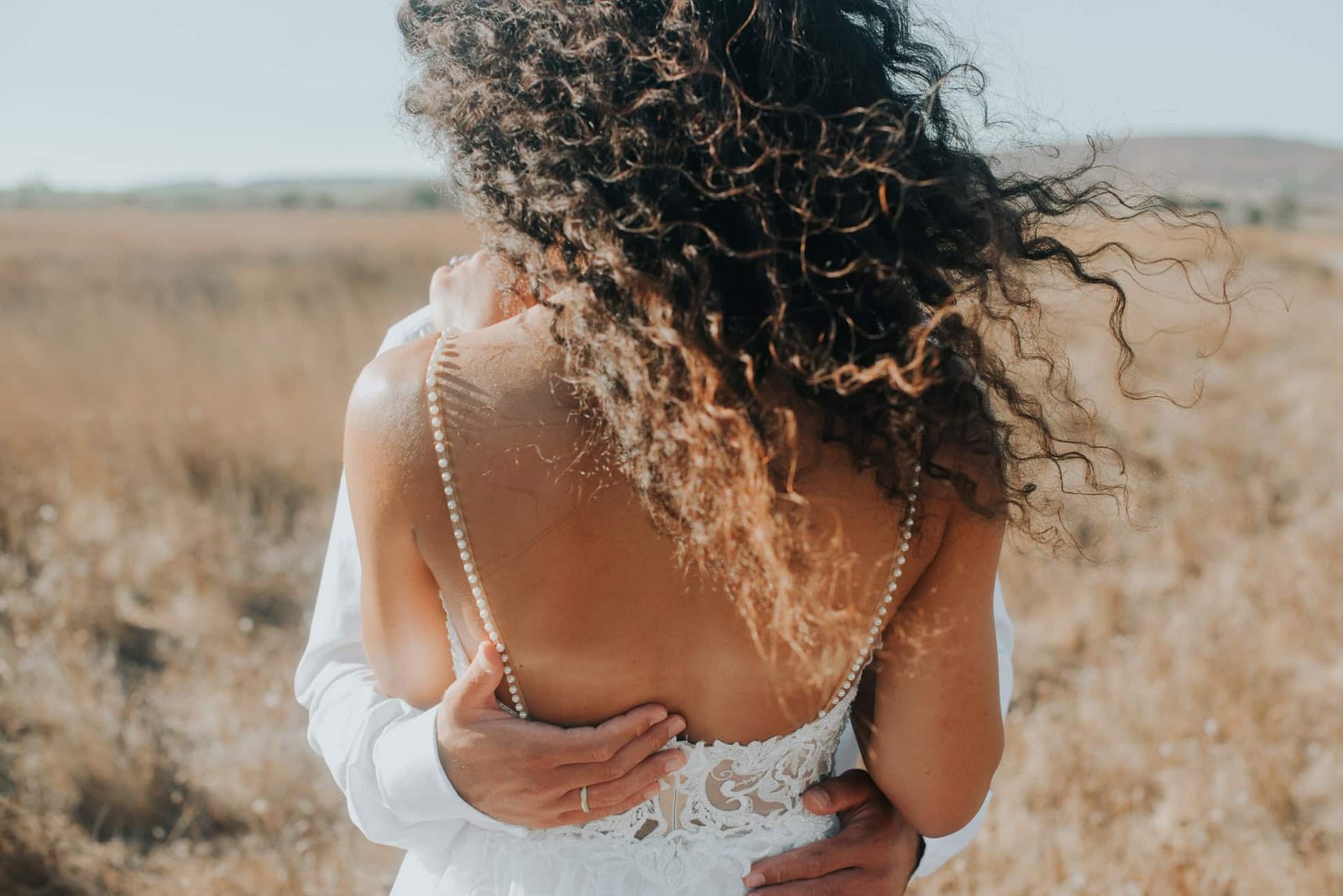 Groom embracing bride from behind showing low back pearl strap wedding dress detail on Greek beach
