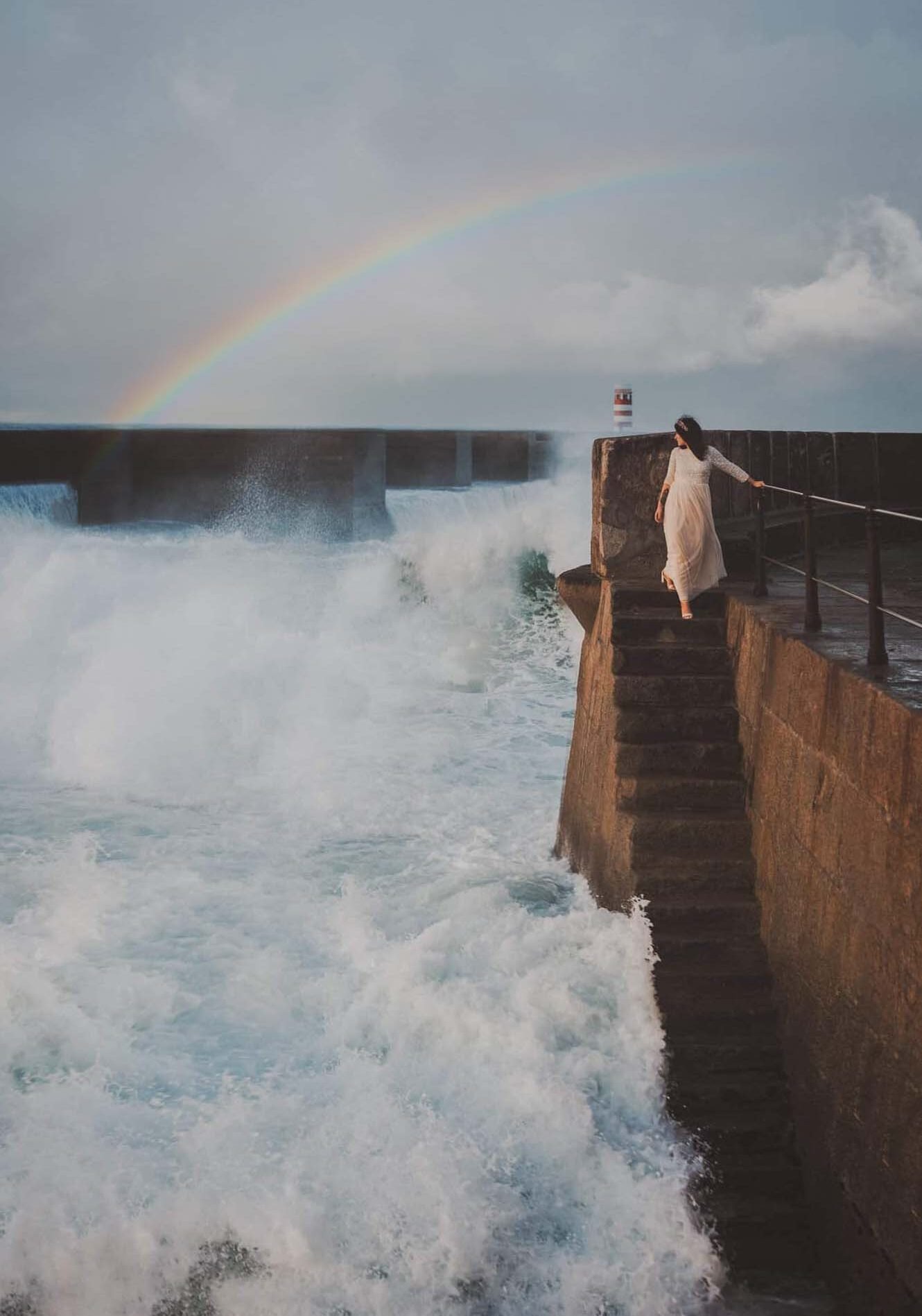 Editorial bride in white lace dress standing on stone pier steps as dramatic waves crash, Foz do Douro Porto Portugal