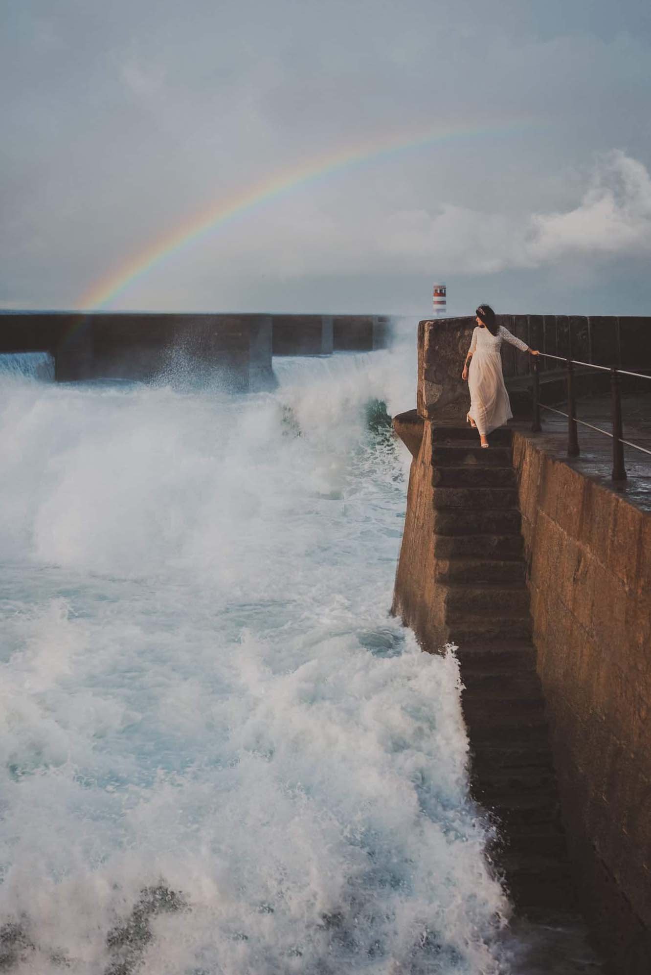 Bride in white lace dress standing on stone pier steps as dramatic waves crash, Foz do Douro Porto Portugal