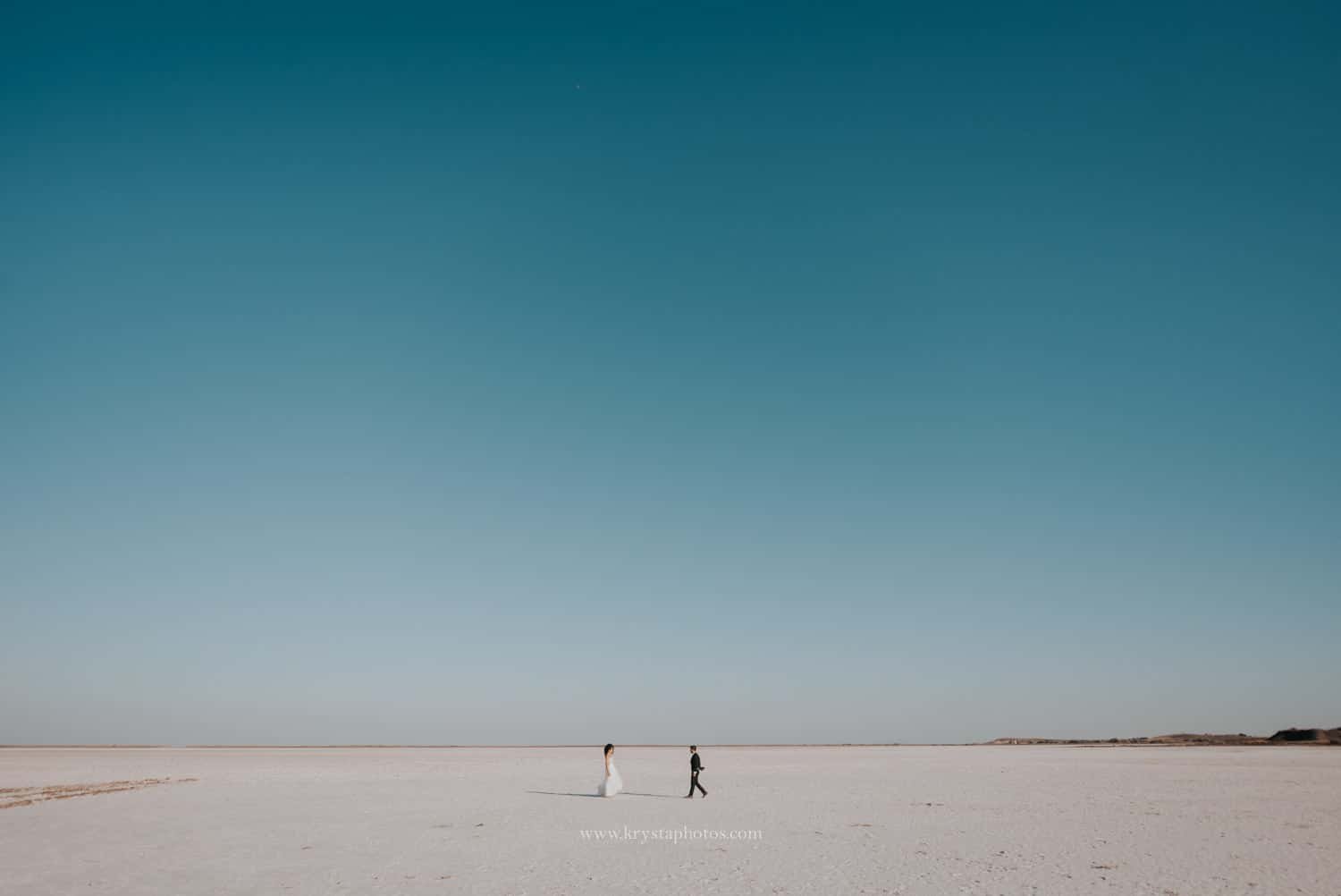 Bride and groom walking across the white salt flats under a clear blue sky in Lemnos, Greece