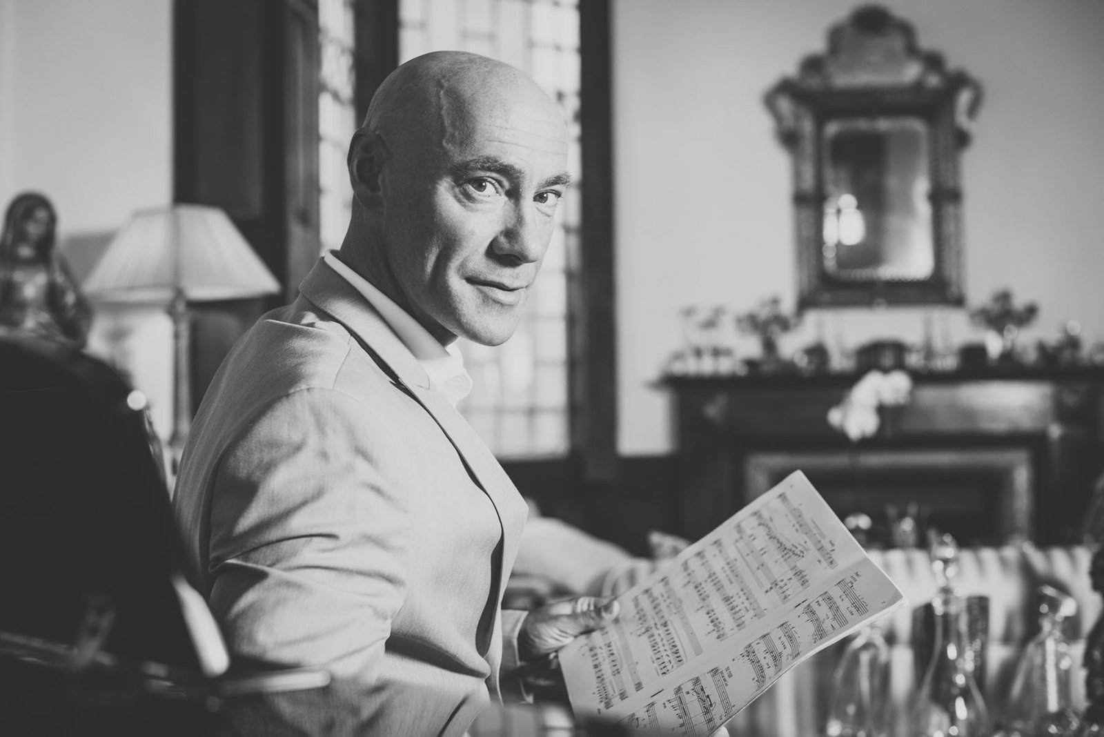 : Bald man in a light suit smiling and glancing at the camera while holding sheet music, seated at a piano in front of an ornate fireplace, black and white portrait