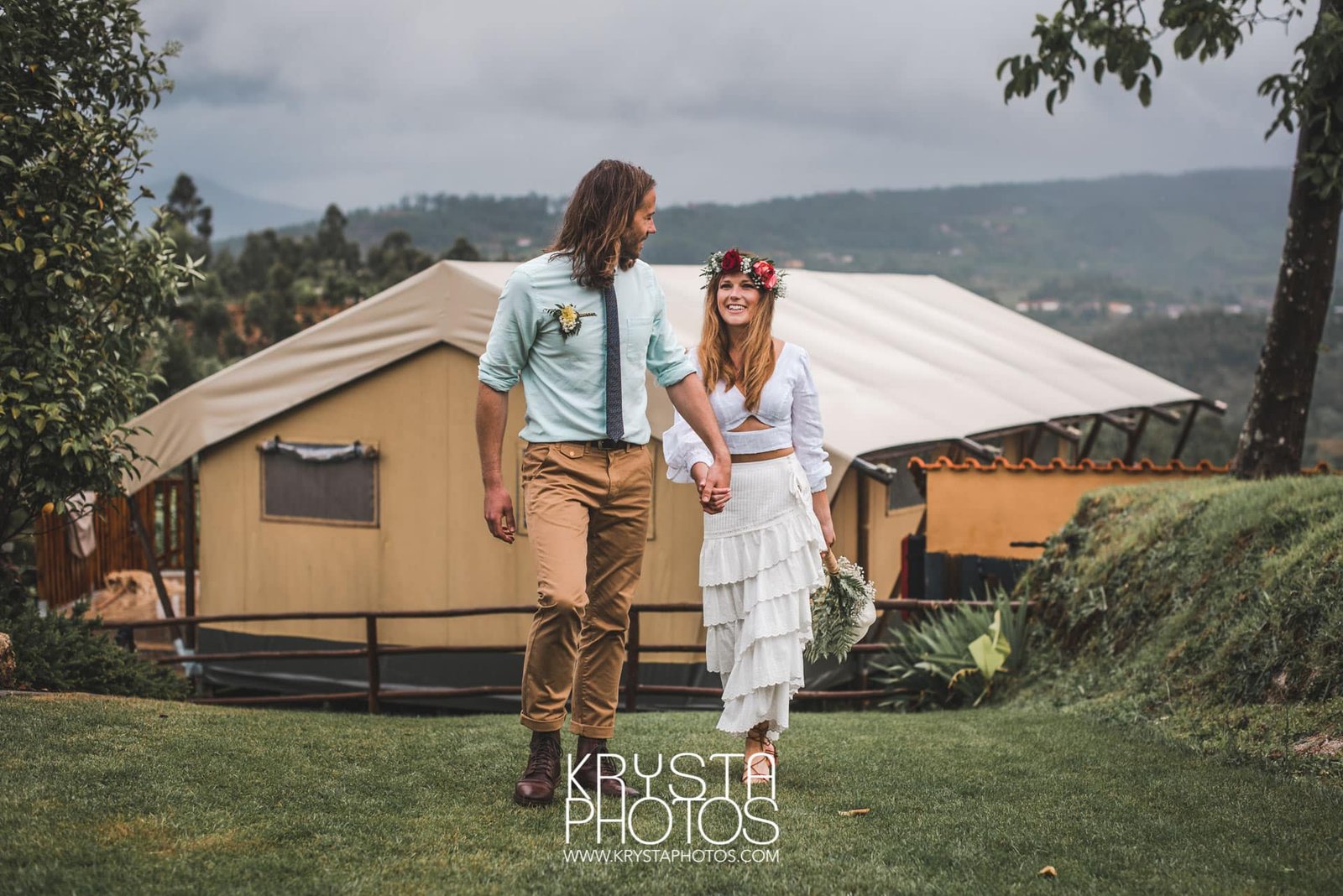 Bride in boho chic dress with flower crown and groom standing together in front of a yellow glamping lodge on their rainy wedding day