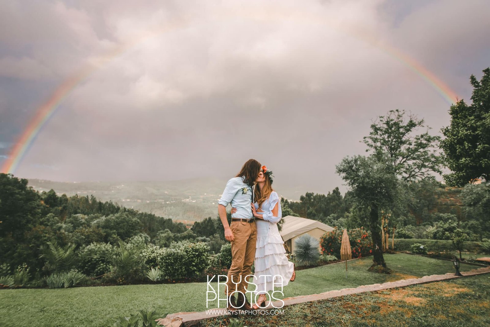Bride in boho chic dress with flower crown standing with groom under a rainbow on their rainy wedding day