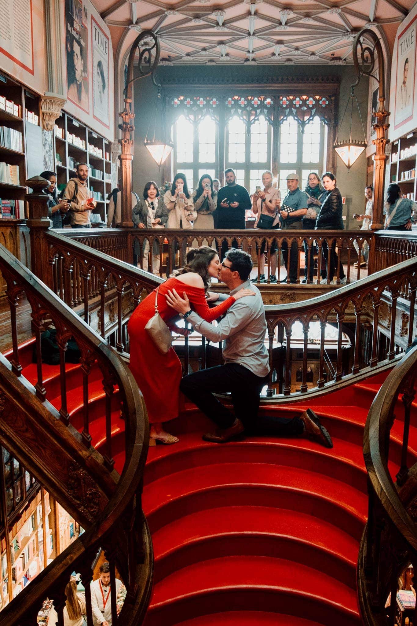 Marriage proposal on the iconic red staircase of Livraria Lello in Porto, Portugal, surrounded by tourists.