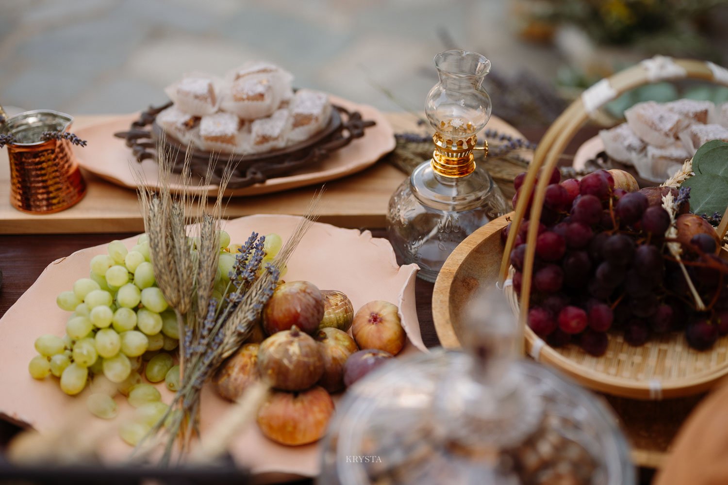 Terracotta plates with green grapes, figs, dried wheat and lavender beside a brass oil lamp and copper briki on a styled table in Lemnos, Greece
