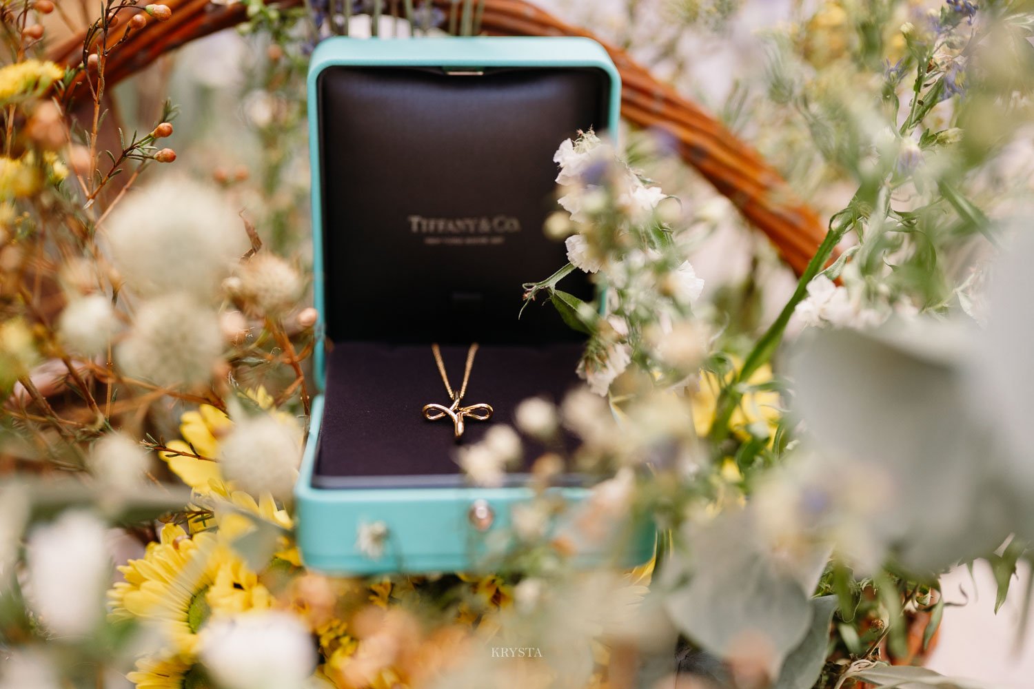 Gold bow cross pendant in a Tiffany & Co. box nestled among yellow and white wildflowers at a celebration in Lemnos, Greece
